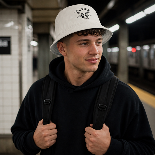 Man wearing white ALTRD bucket hat with logo, black hoodie, and backpack at subway station