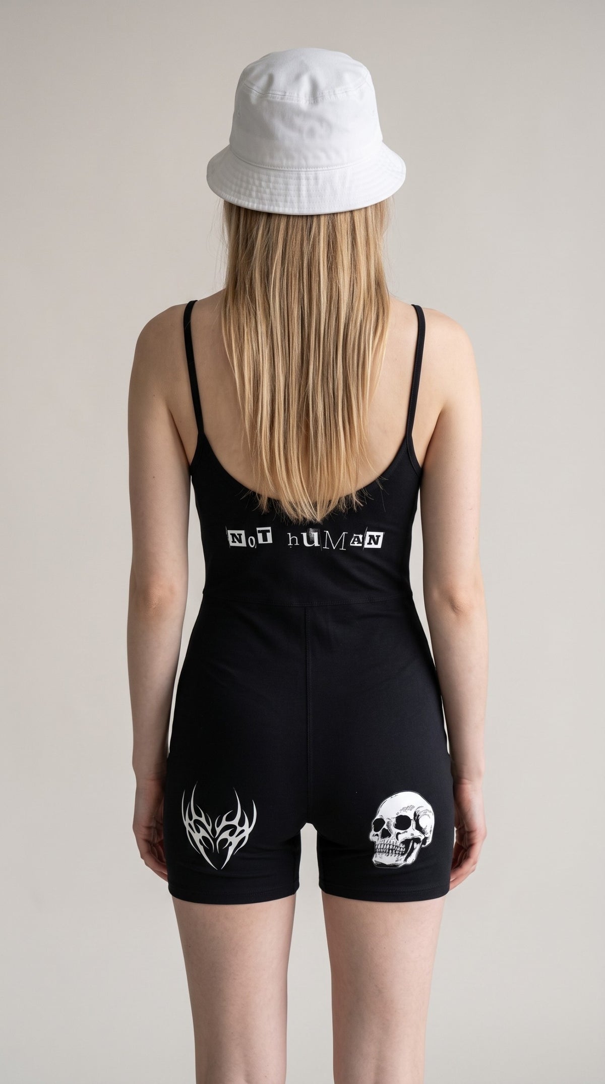 Back view of a woman wearing an ALTRD Apparel black yoga romper with NOT HUMAN text and skull graphic, white bucket hat